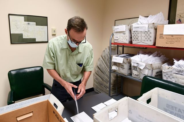 A man wearing a mask looks at absentee ballots in an office room full of boxes of absentee ballots